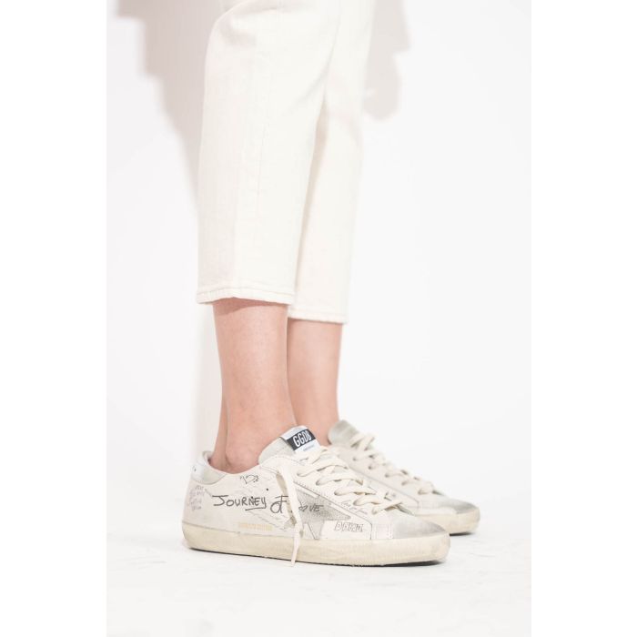 Sneakers en cuir blanc Graffiti écritures et dessins noirs arrière en cuir argent Golden Goose femme. Edition limitée. Portées de profil avec un jean Boy Straight écru stretch R13.