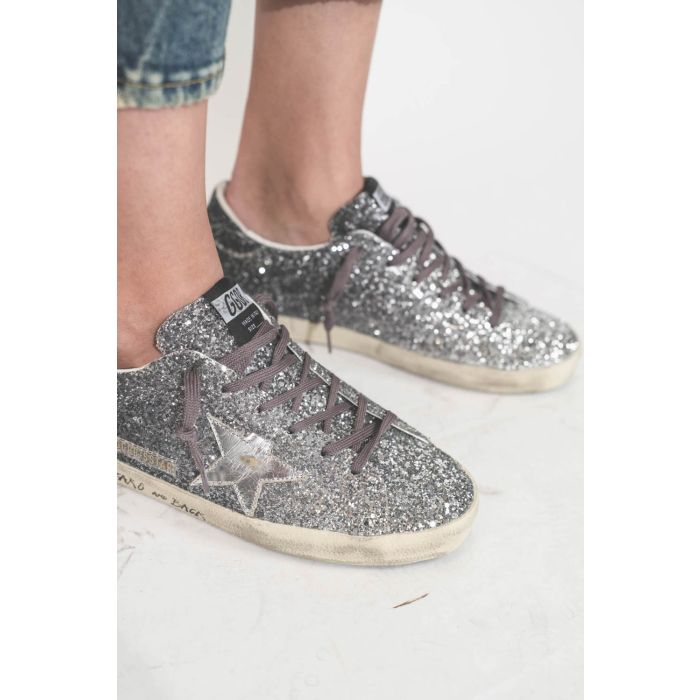 Sneakers entièrement recouvertes de paillettes argentées et noires, étoile en cuir lisse tague écritures noires sur la semelle droite beige "LOVE You To The Star And Back"  Super-star Golden Goose femme. Portées de profil avec un jean Boy Straight bleu R1