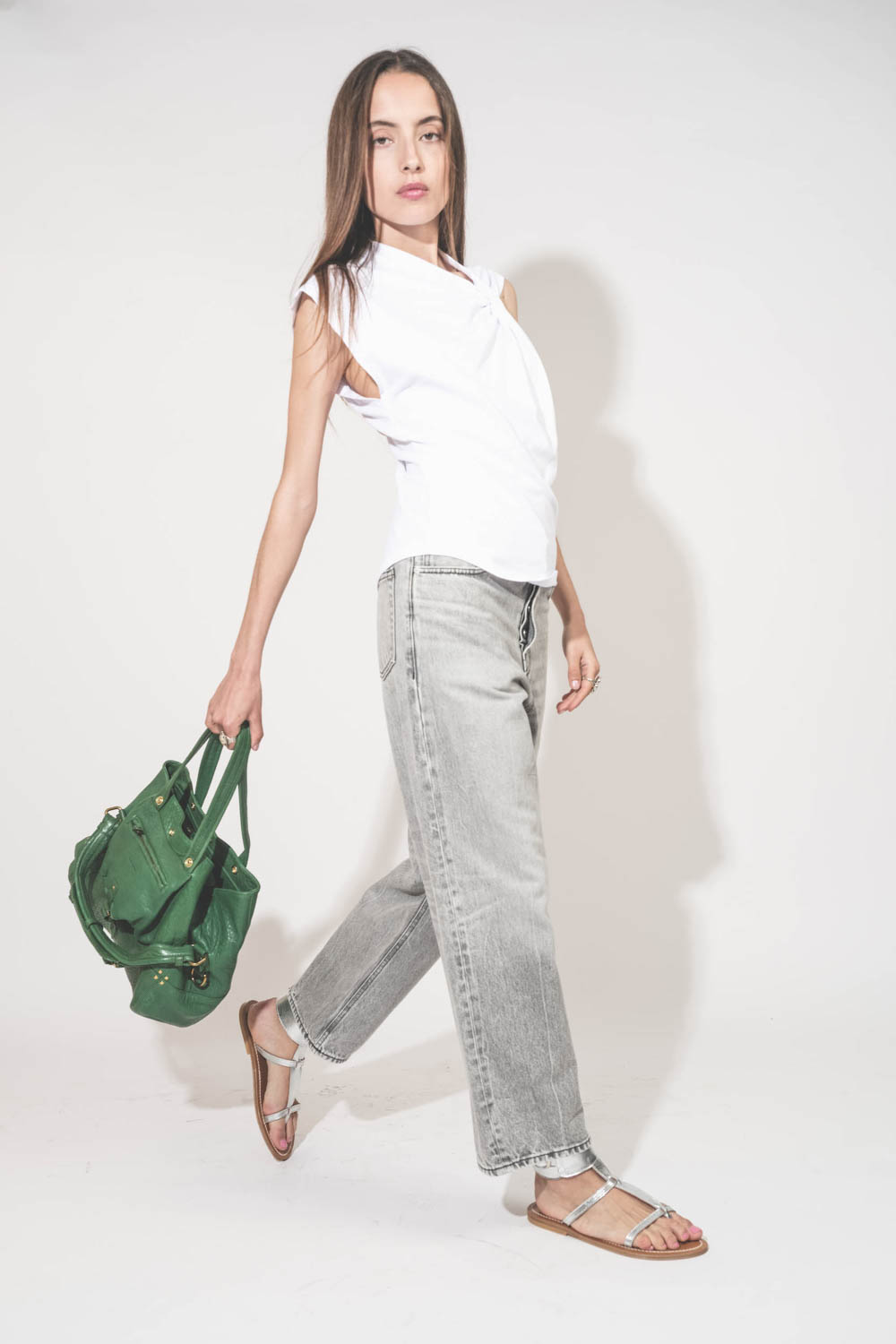Tee-shirt sans manches noué sur le devant de l'épaule asymétrique en jersey blanc Nayda Marant Etoile. Porté de profil avec une jean large gris Betty Haikure et des sandales plates en cuir argent Caravelle K.Jacques St-Tropez. Sac cabas porté main en cuir