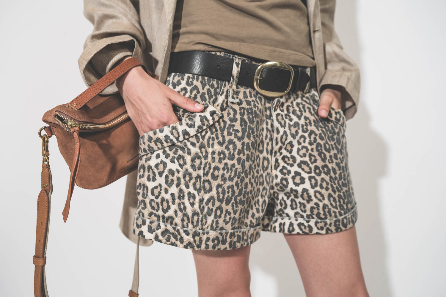 Pochette en cuir suédé caramel Lucky Jérôme Dreyfuss portée main avec une bandoulière sangle large écru Jérôme Dreyfuss. Alyssa porte une veste Ragged Oversized beige en lin R13 avec un short en toile léopard Pocket R13. Ceinture en cuir noir Dara Isabel 