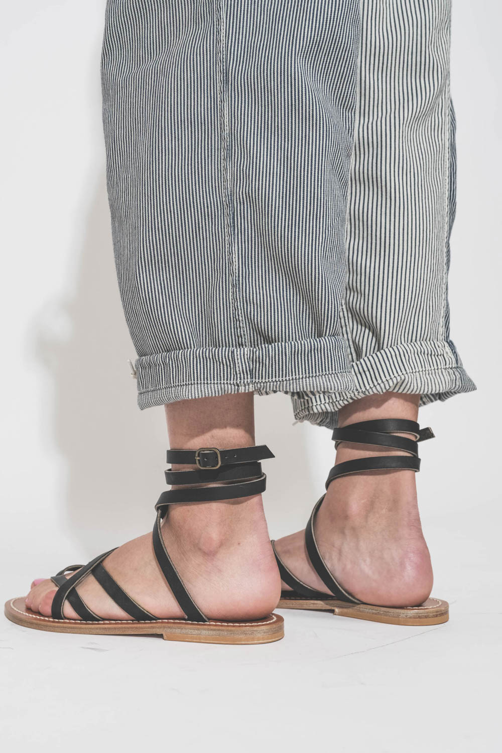 Sandales plates en cuir noir Zenobie K.Jacques Saint-Tropez. Porté de dos avec un pantalon à rayures Cargo Railroad R13 denim.