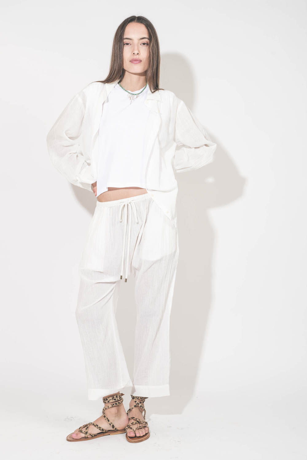 Sandales plates avec brides Zenobie K.Jacques St-Tropez en cuir "pony" léopard. Porté de face avec un chemise en voile de coton blanc et un pantalon assorti en voile de coton blanc The Great. Tee-shirt sans manches blanc The Peak Shoulder The Great.