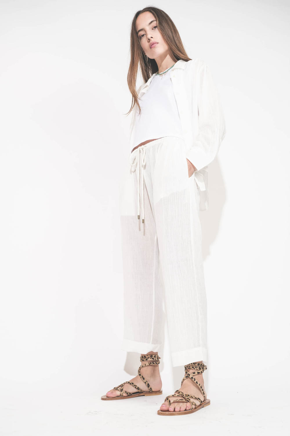Pantalon Sarouel en voile de coton blanc Reef The Great. Porté de profil avec une chemise manches longues en voile de coton blanc et un tee-shirt sans manches Peak Shoulder The Great. Sandales en cuir pony léopard Zenobie K.Jacques St-Tropez. Collier turq
