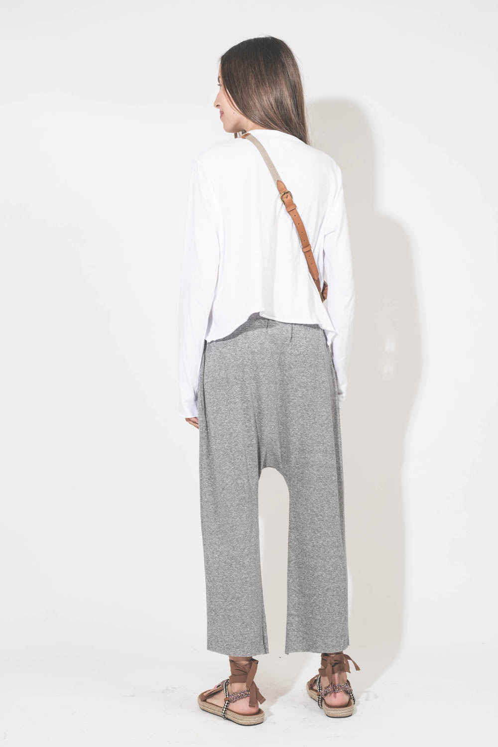 Pantalon sarouel court en jersey gris chiné The Great. Porté de dos avec une tee-shirt court manches longues blanc et des sandales en cordes tressées et ruban beige Gypsa Isabel Marant. Pochette Lucky en cuir suédé caramel portée crossbody avec une sangle