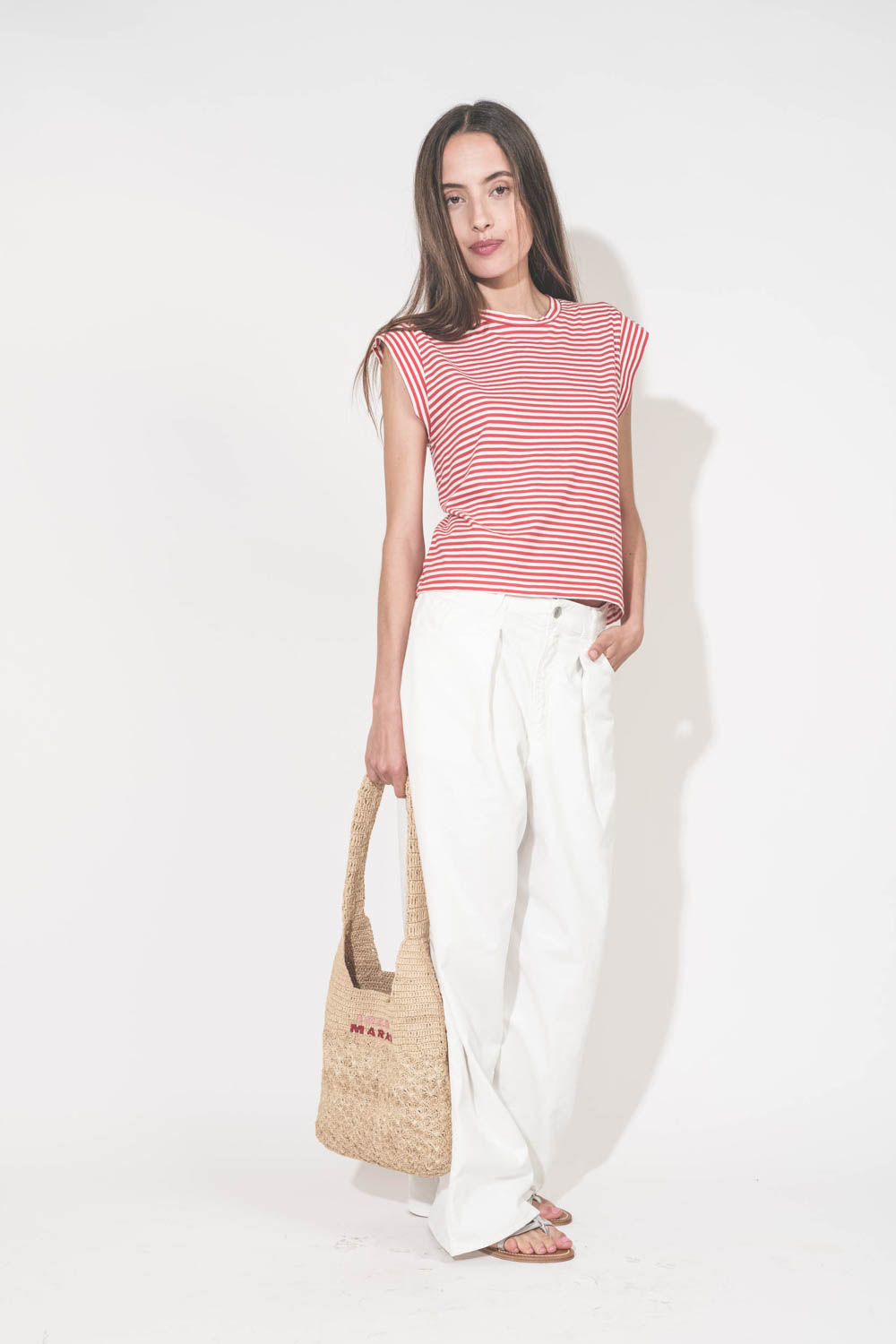 Tee-shirt manches courtes raccourcies en coton rayé rouge et blanc Peak Shoulder The Great. Porté de face avec un jean large blanc Georgia Denimist et des sandales en cuir argent Caravelle K.Jacques St-Tropez. Panier en osier naturel Praia Isabel Marant.