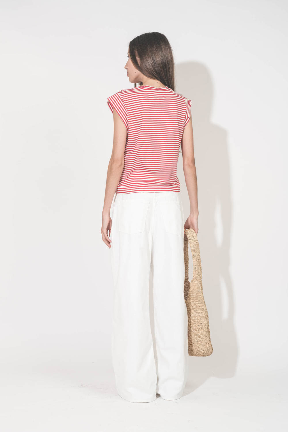 Tee-shirt manches courtes raccourcies en coton rayé rouge et blanc Peak Shoulder The Great. Porté de dos avec un jean large blanc Georgia Denimist et des sandales en cuir argent Caravelle K.Jacques St-Tropez. Panier en osier naturel Praia Isabel Marant.